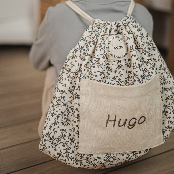 Child wearing a floral drawstring bag with 'Hugo' on a wooden floor